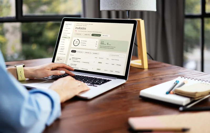 Una planner trabaja en su laptop sobre un escritorio de madera; en la pantalla, el panel de MONÉ con la lista de invitados de una boda